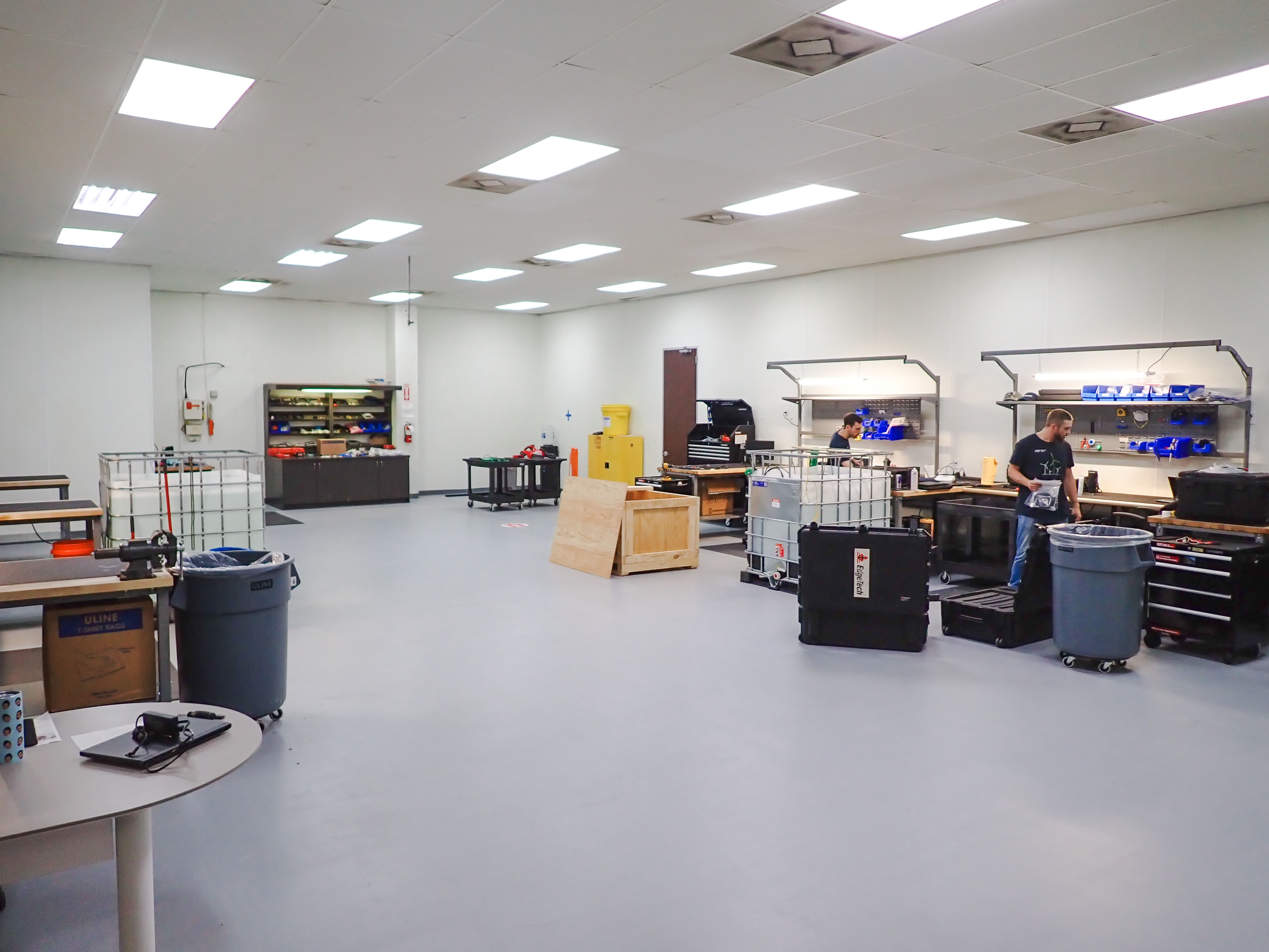 Two men work in a brightly lit workshop with multiple workstations, tools, and storage units.