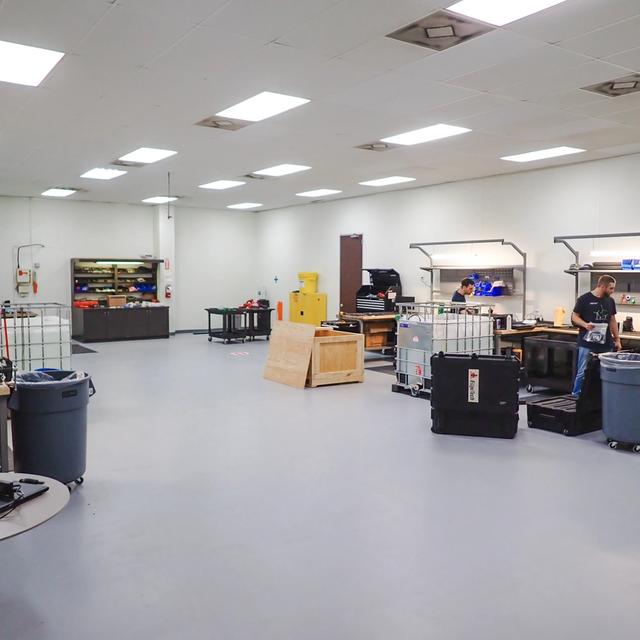 Two men work in a brightly lit workshop with multiple workstations, tools, and storage units.