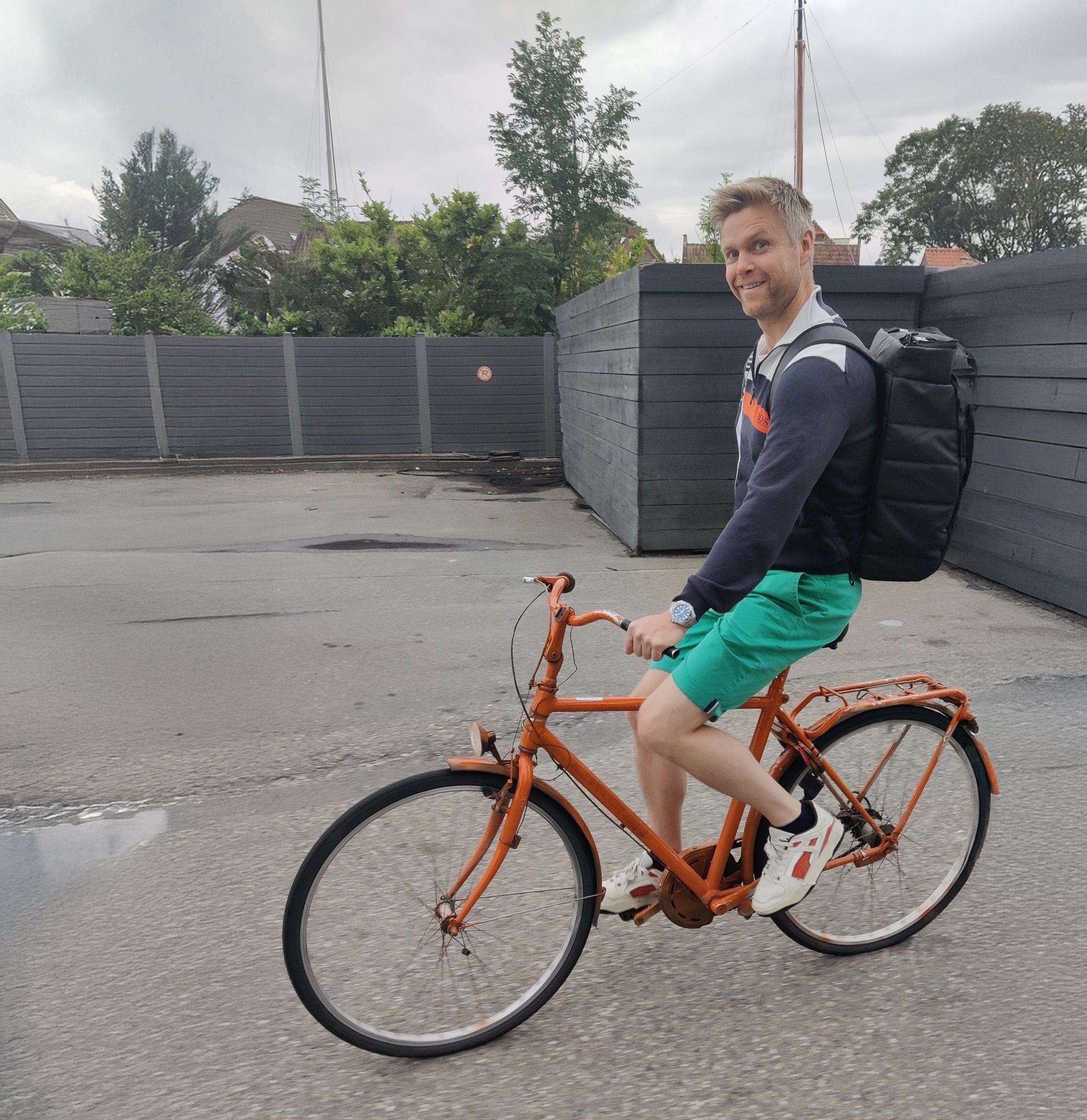 A smiling man with a black backpack rides an orange bicycle.