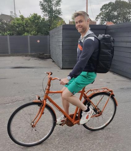 A smiling man with a black backpack rides an orange bicycle.