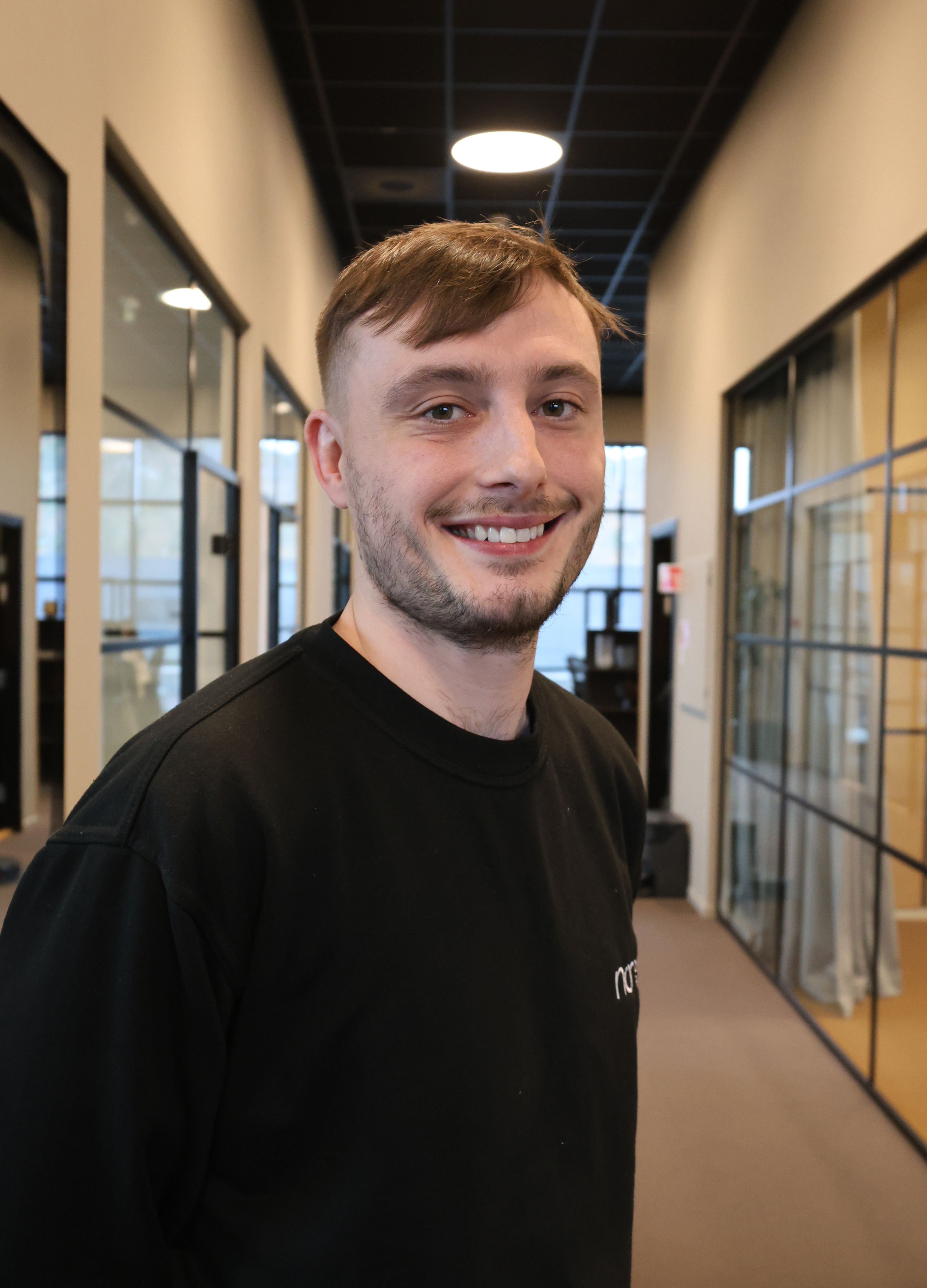Young man smiling in a black sweatshirt in an office hallway.