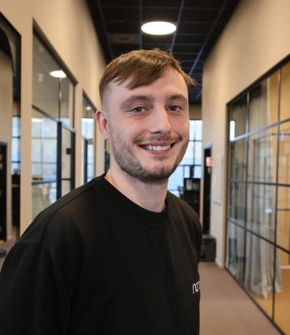 Young man smiling in a black sweatshirt in an office hallway.