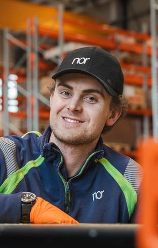 A smiling man wearing a black "nør" cap and blue work jacket with an orange glove, in a warehouse.