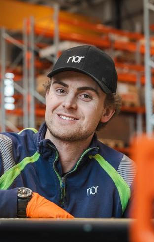 A smiling young man in a "nor" cap and work jacket with orange gloves in an industrial setting.