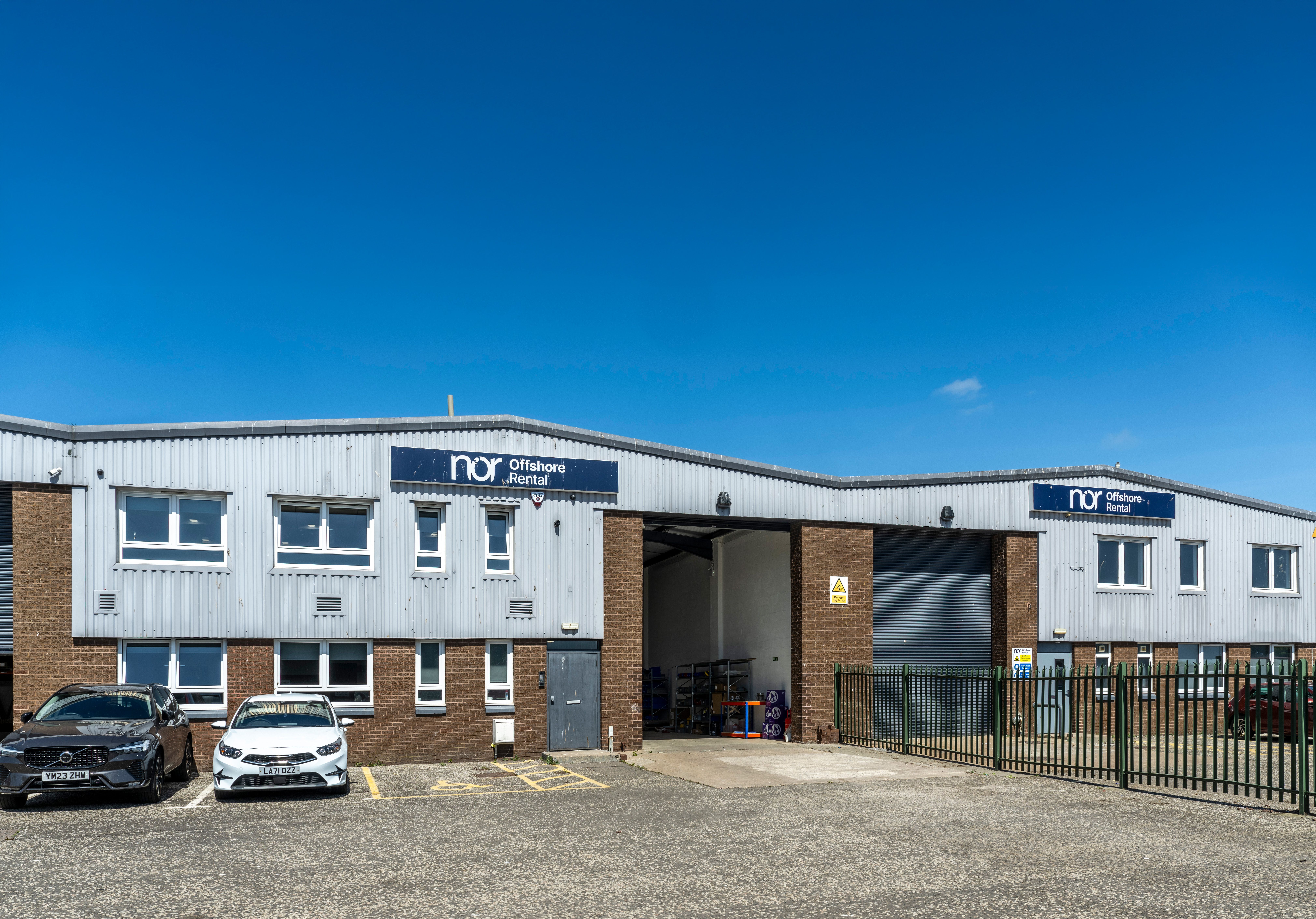 A gray industrial building with a "nor Offshore Rental" sign, two cars parked outside, and a clear blue sky.