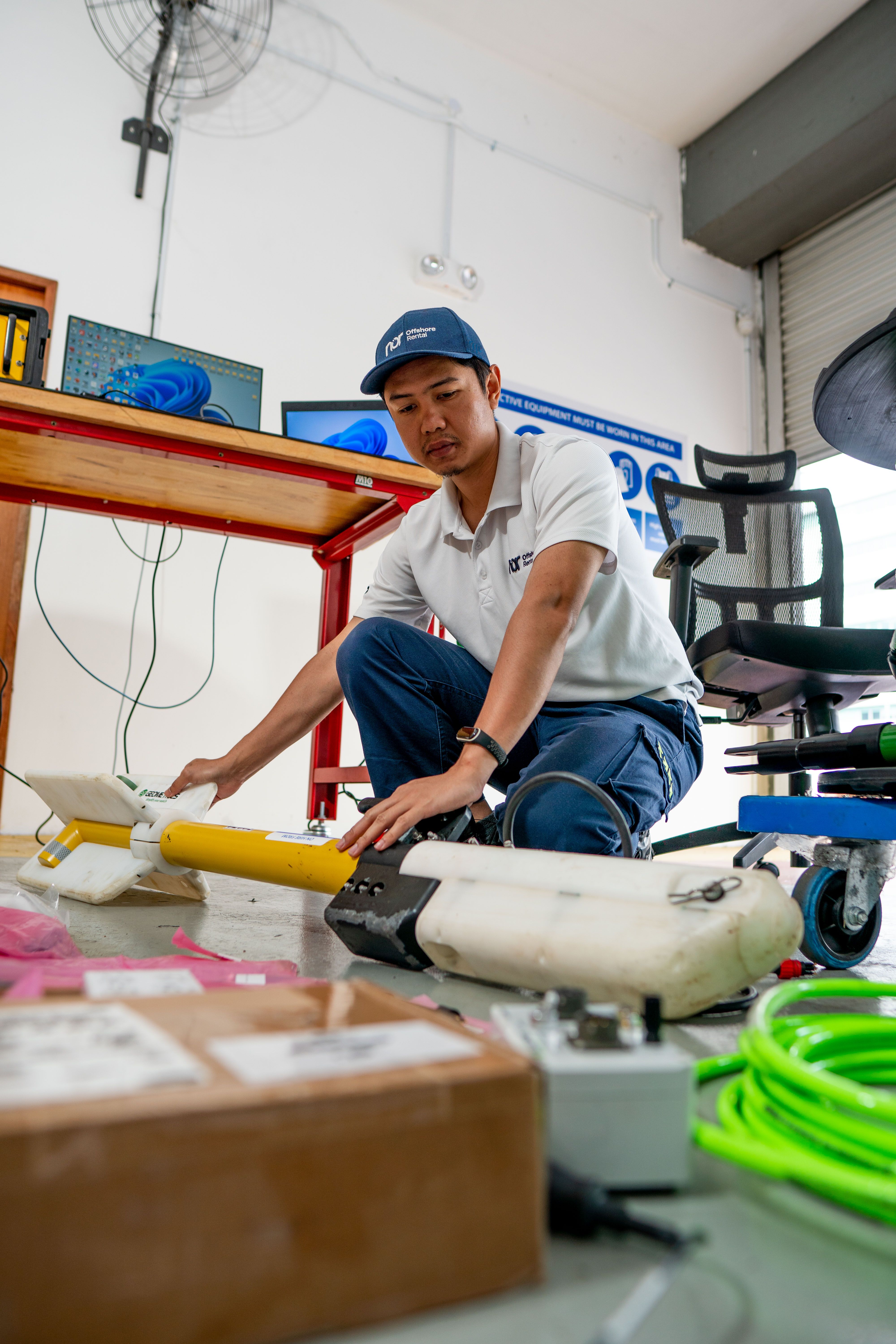 A man in a Nor Offshore Services uniform crouches, working on yellow and white oceanographic equipment on the floor.