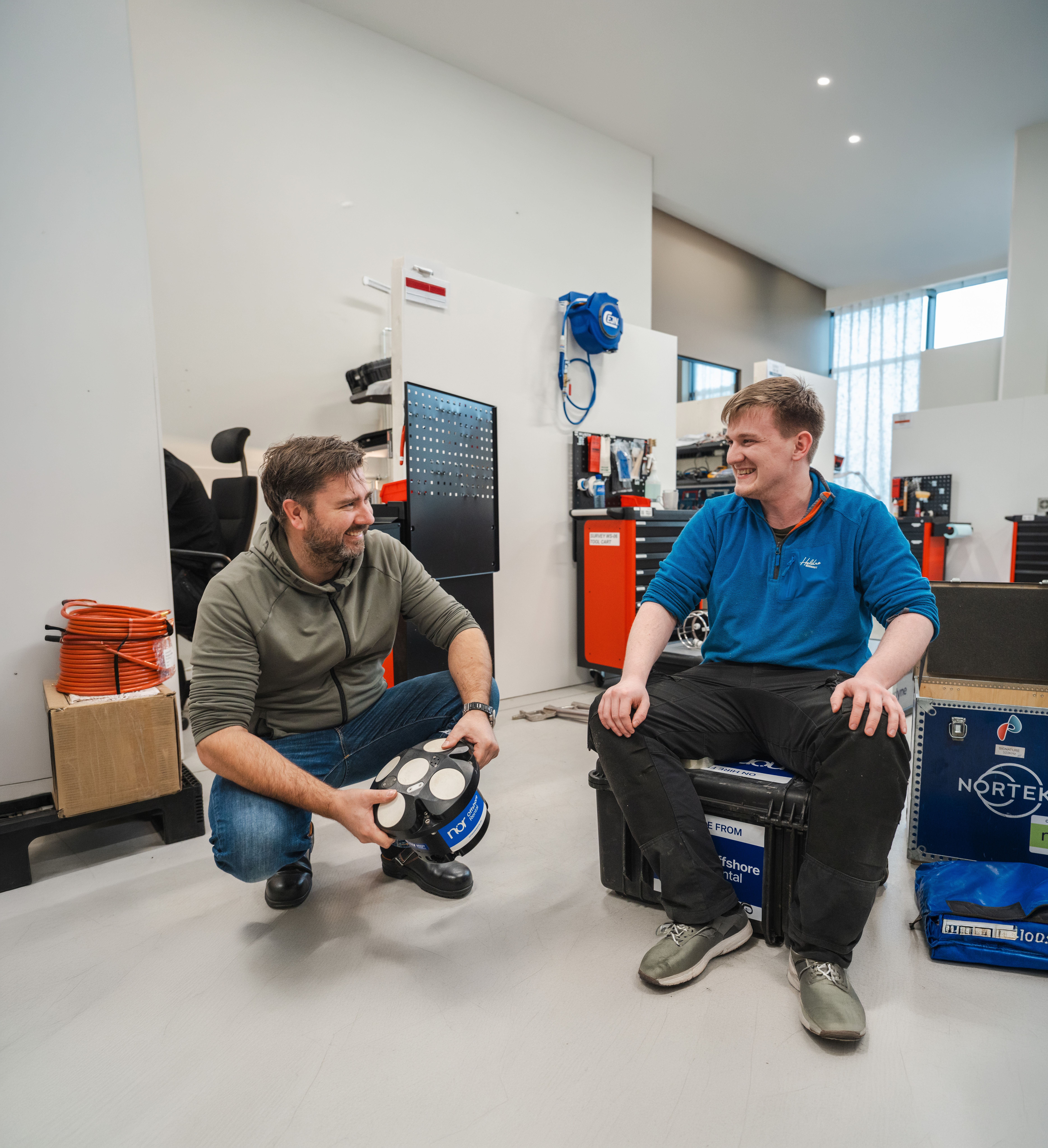 Two men smiling in a workshop, one crouching and holding a Nortek sensor, the other sitting on a protective case.