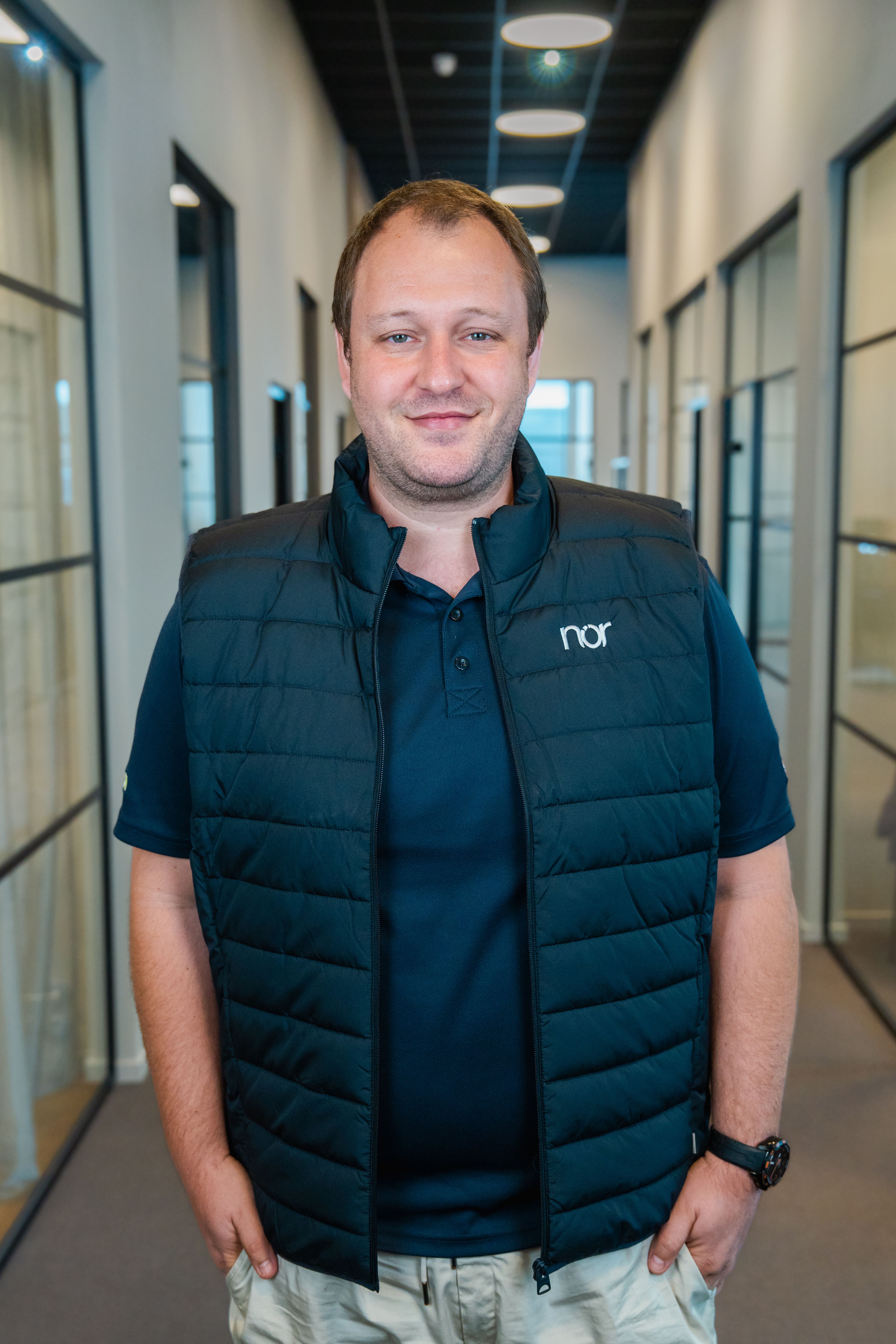 A smiling man in a dark blue vest with "nor" logo and light pants stands in an office hallway.
