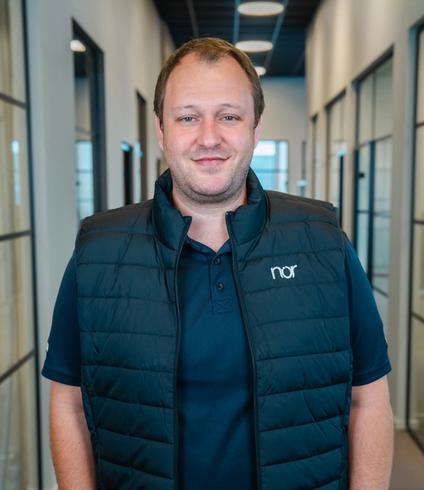 A smiling man in a dark blue vest with "nor" logo and light pants stands in an office hallway.