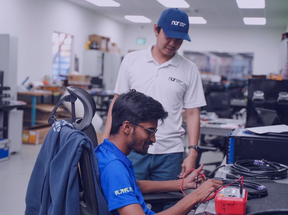 Two men in a lab, one testing cables with a multimeter while the other observes.