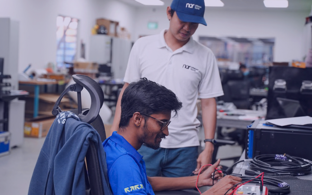 Two men in a lab, one testing cables with a multimeter while the other observes.