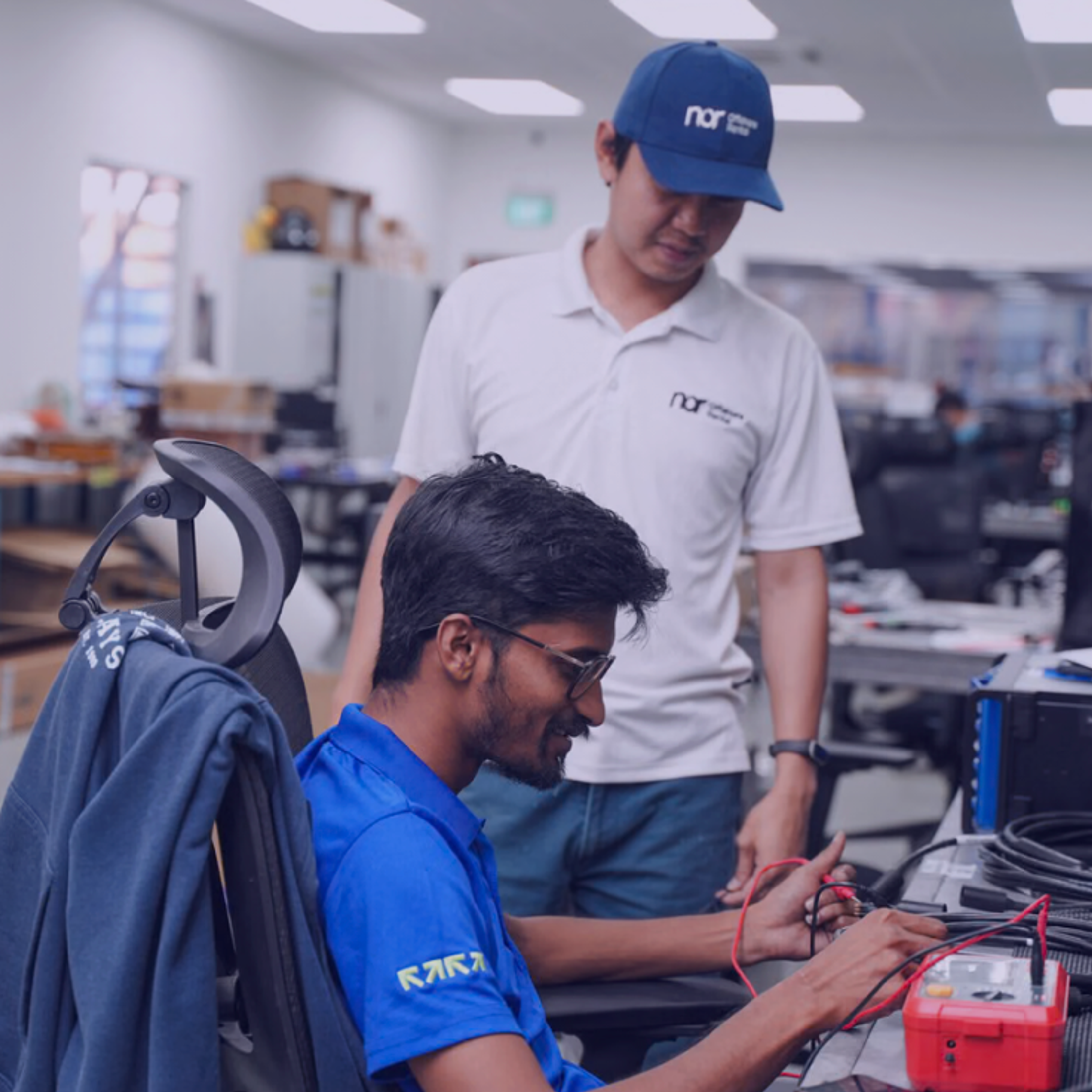 Two men in a lab, one testing cables with a multimeter while the other observes.