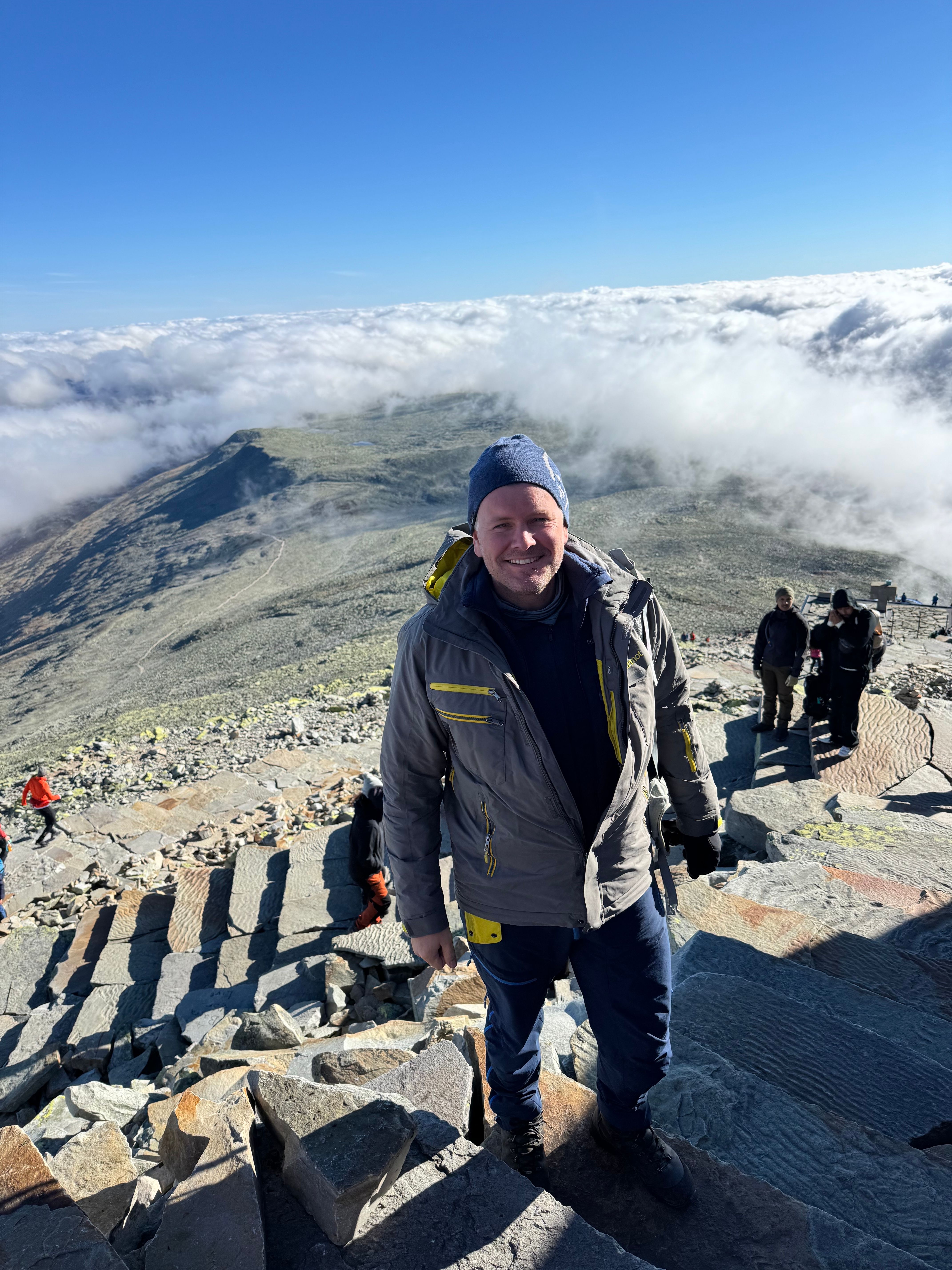 A man smiles on a rocky mountaintop overlooking a vast cloudscape and distant peaks under a blue sky.