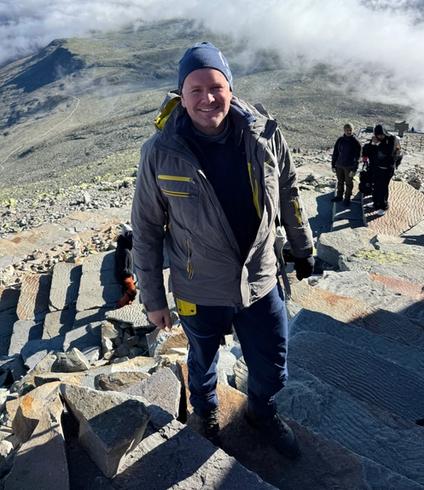 A man smiles on a rocky mountaintop overlooking a vast cloudscape and distant peaks under a blue sky.