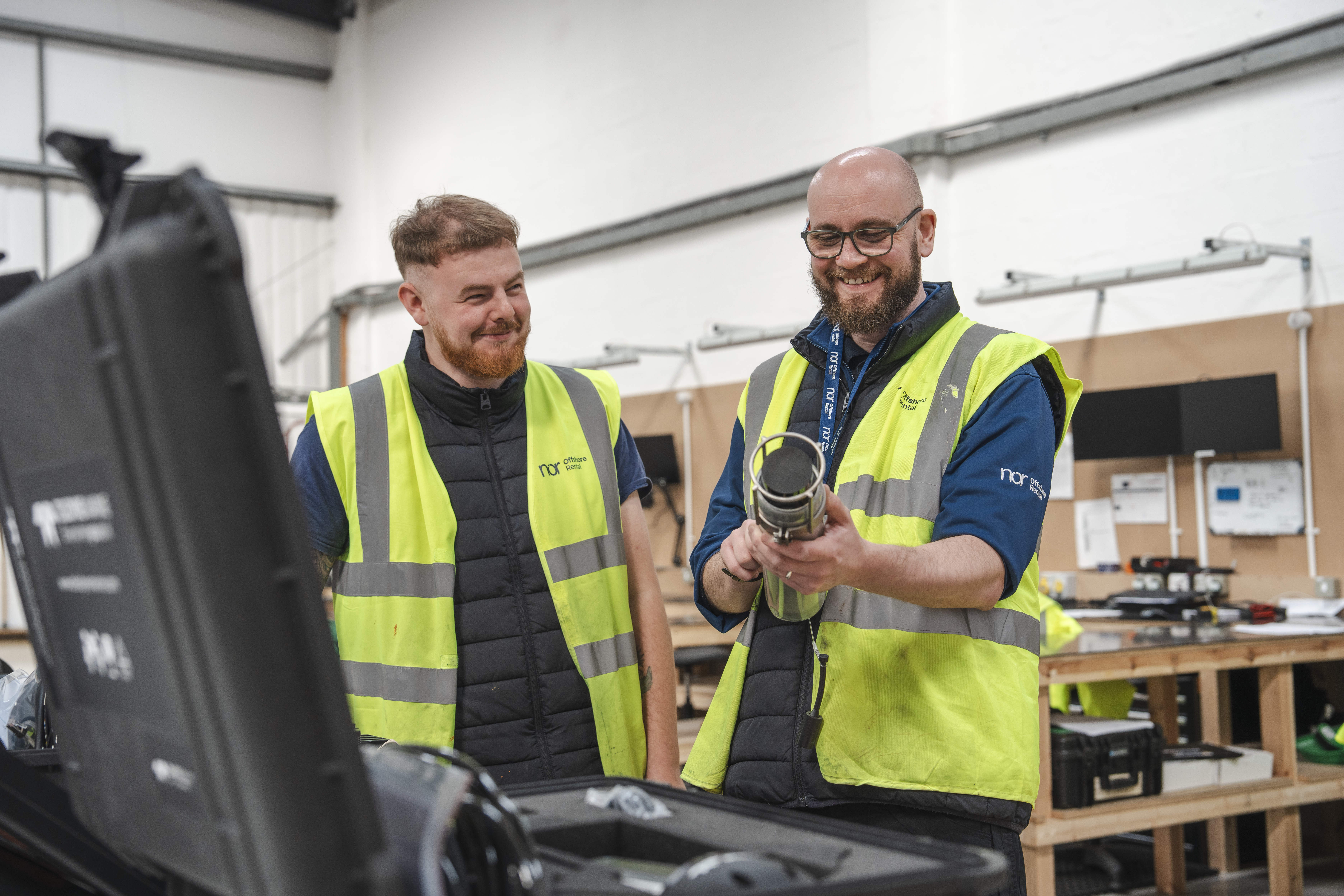 Two smiling men in high-visibility vests examine a cylindrical device in a workshop.