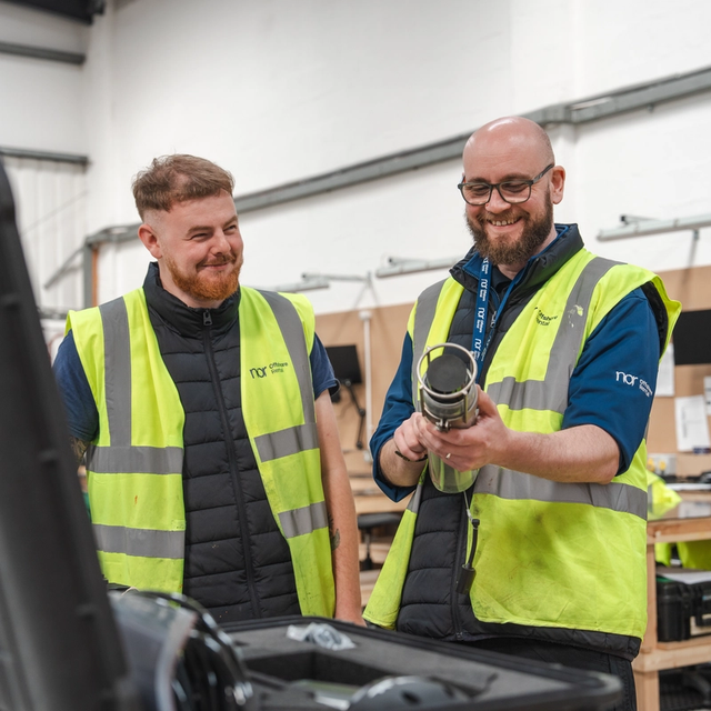 Two smiling men in high-visibility vests examine a cylindrical device in a workshop.
