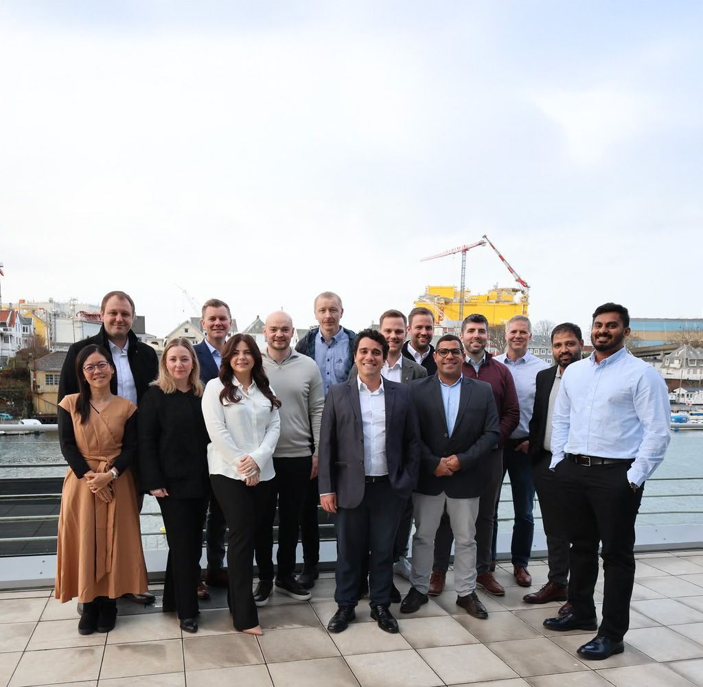 A group of 15 smiling professionals standing outdoors by a waterfront with an industrial backdrop.