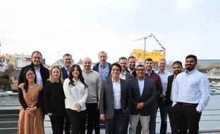 A group of 15 smiling professionals standing outdoors by a waterfront with an industrial backdrop.