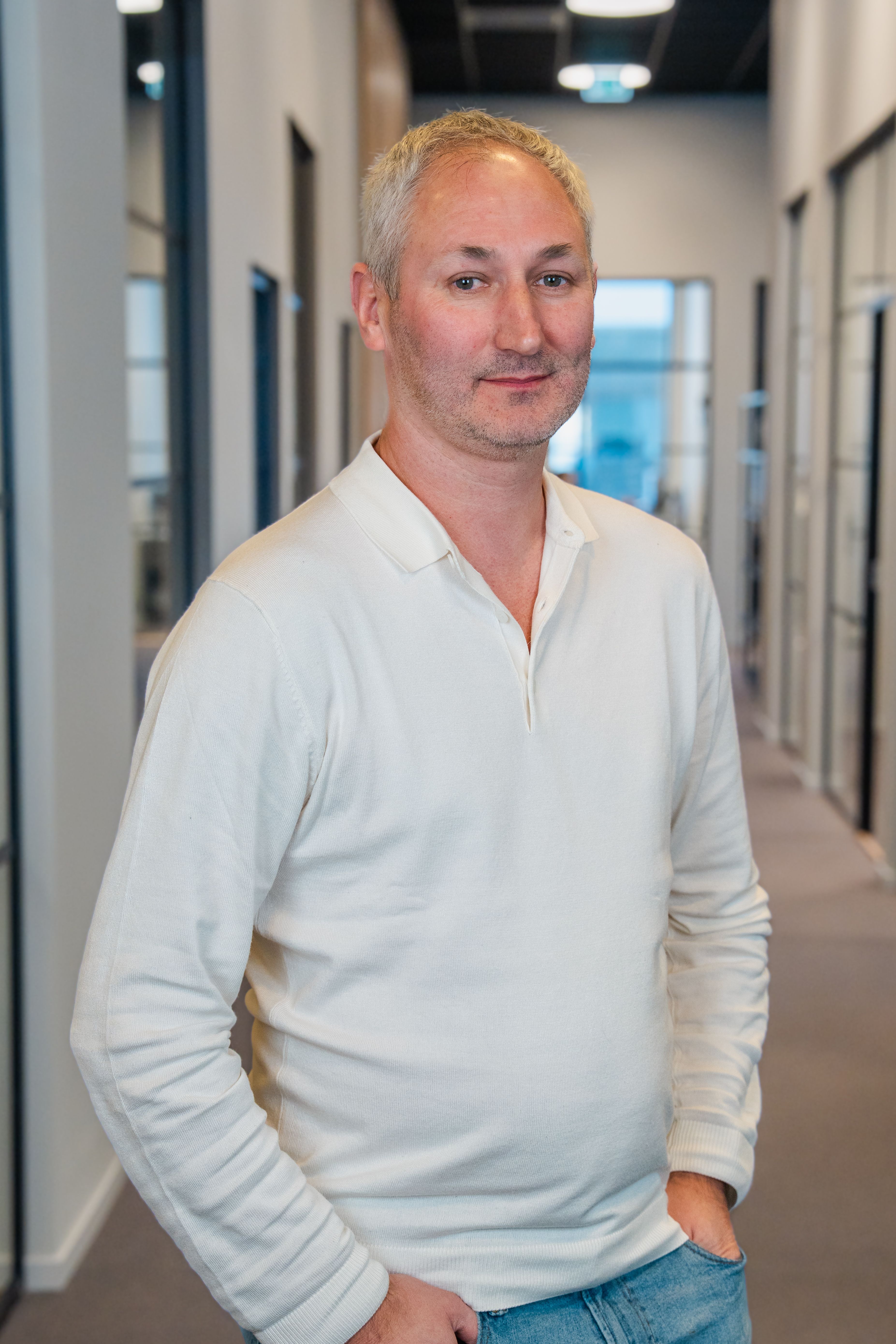 A man with gray hair wearing a cream long-sleeve polo shirt stands in an office hallway.