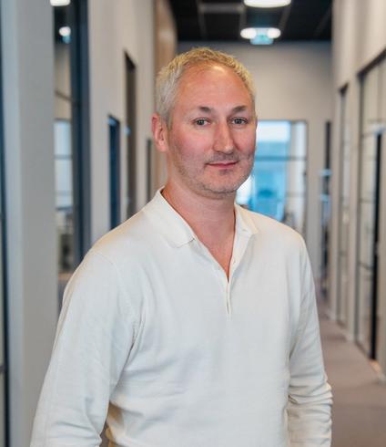 A man with gray hair wearing a cream long-sleeve polo shirt stands in an office hallway.