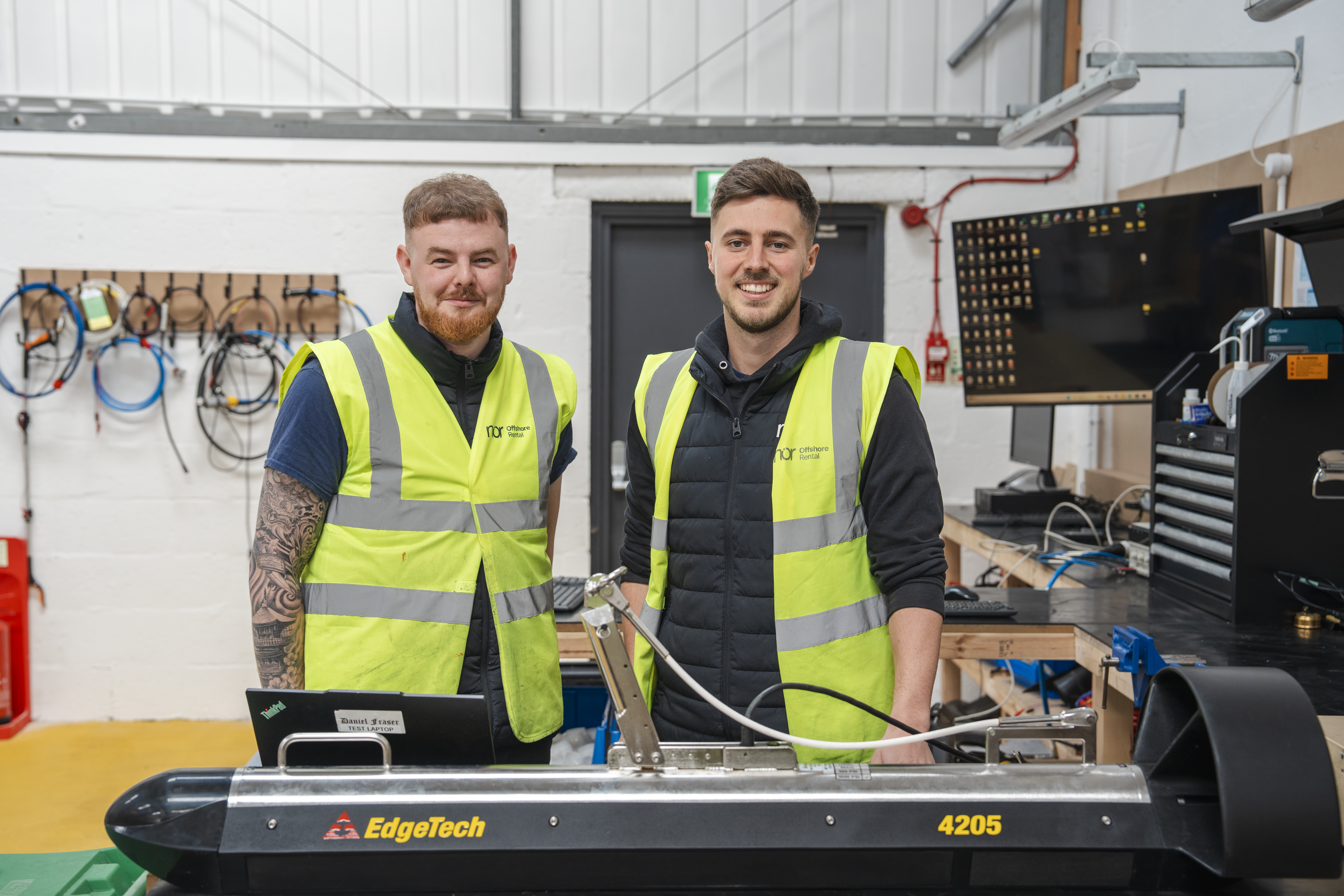 Two smiling men in high-visibility vests stand in a workshop with an EdgeTech 4205 device.
