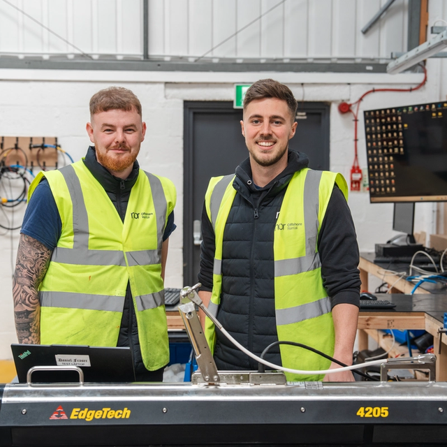 Two smiling men in high-visibility vests stand in a workshop with an EdgeTech 4205 device.