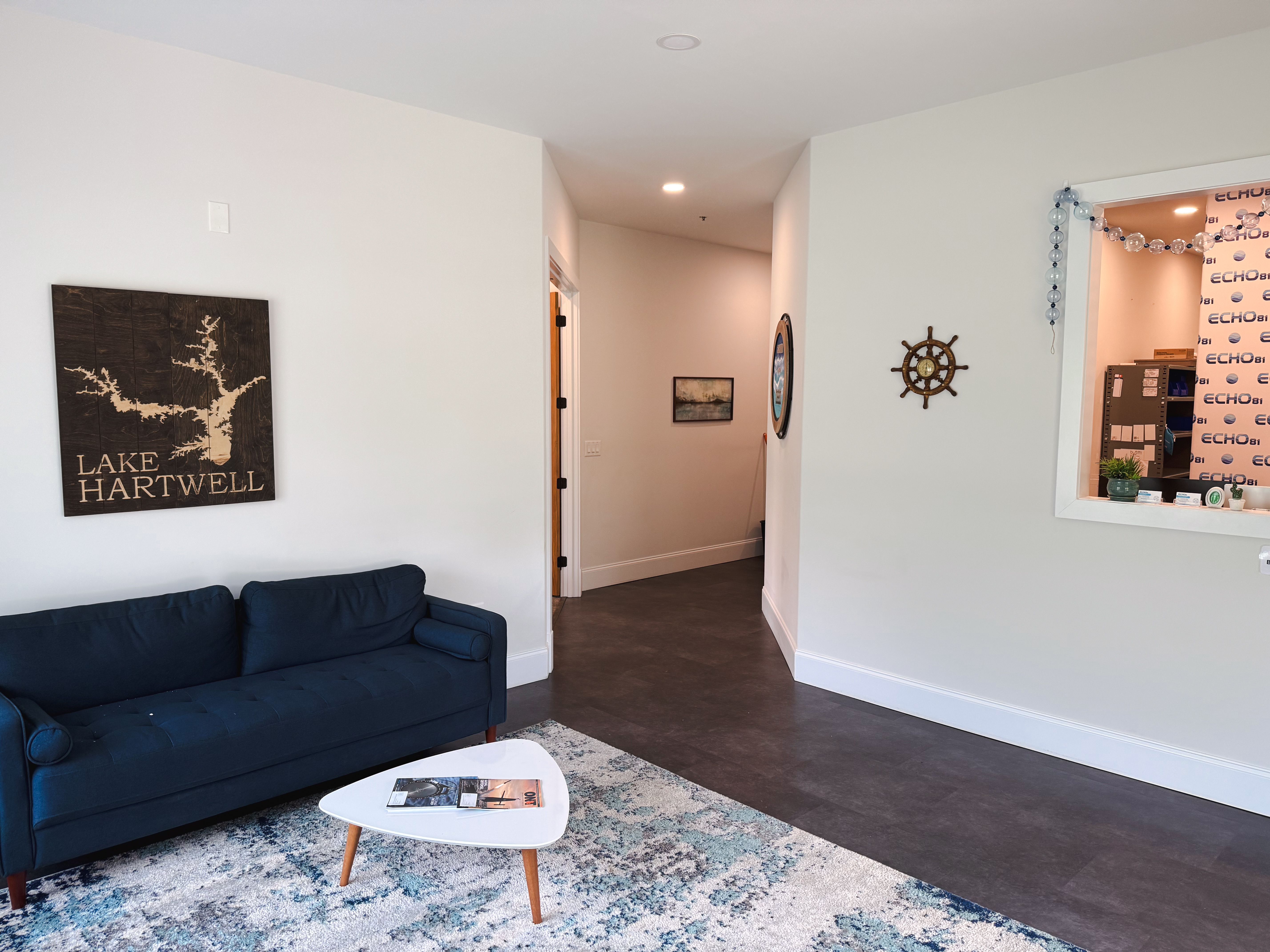 A waiting area with a blue sofa, Lake Hartwell map, ship's wheel decor, and a window to a reception desk.