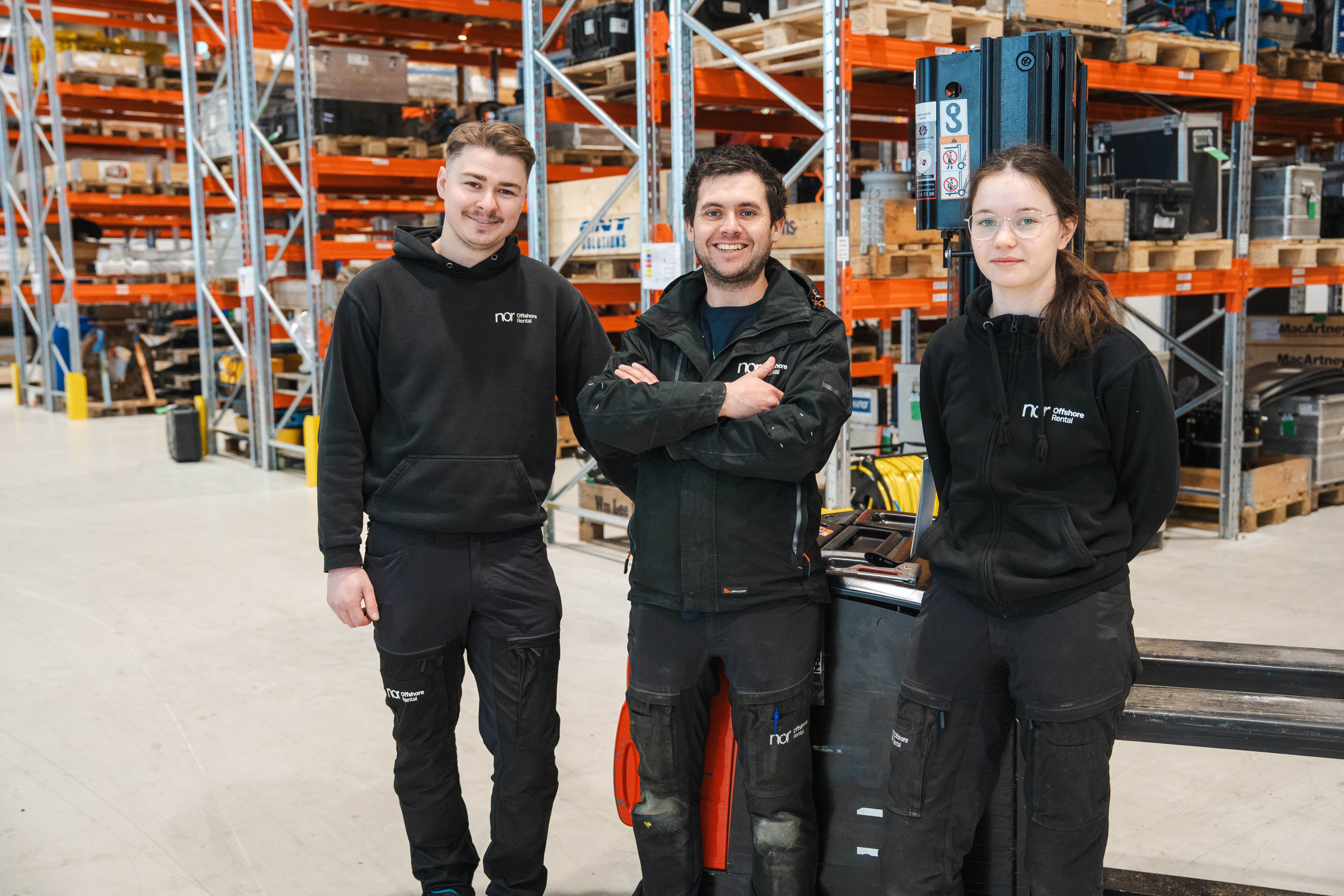 Three people in black workwear smiling in a warehouse.