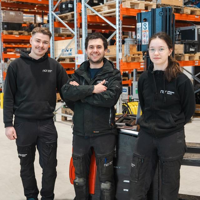 Three people in black workwear smiling in a warehouse.