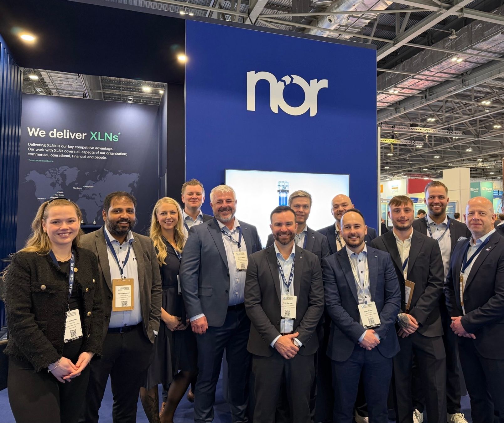 A smiling team stands in front of a blue "nor" booth at a trade show, with "We deliver XLNs" visible on a display.