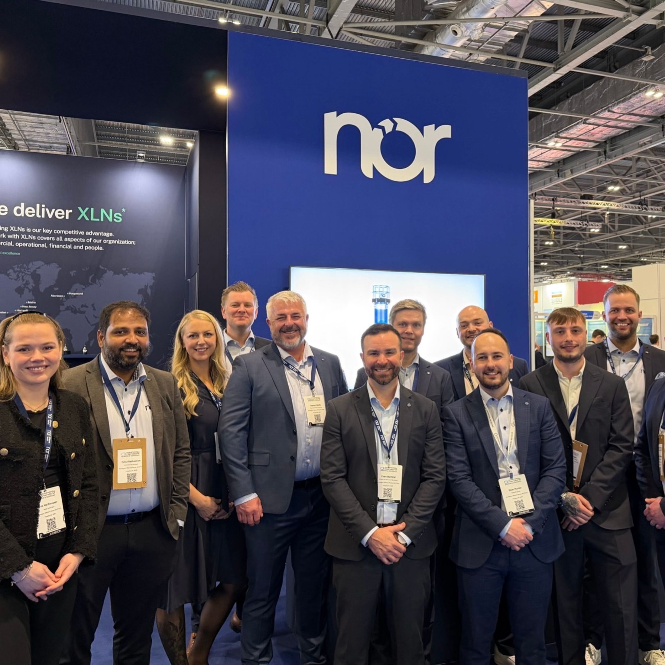 A smiling team stands in front of a blue "nor" booth at a trade show, with "We deliver XLNs" visible on a display.