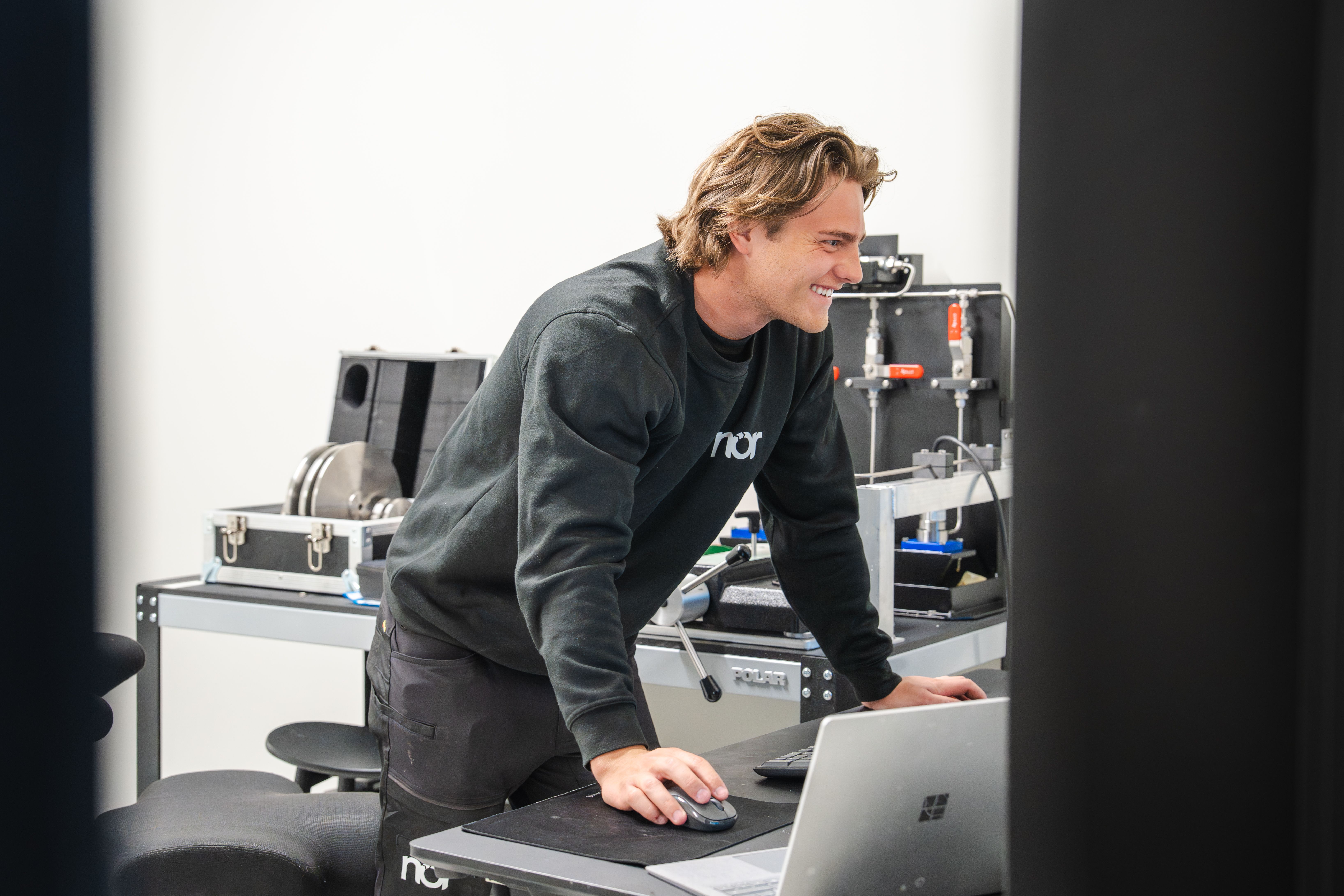 A smiling man in a black "nor" sweatshirt works on a laptop in calibration lab.