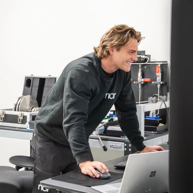 A smiling man in a black "nor" sweatshirt works on a laptop in calibration lab.