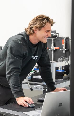 A smiling man in a black "nor" sweatshirt uses a computer mouse at a desk with lab equipment in the background.