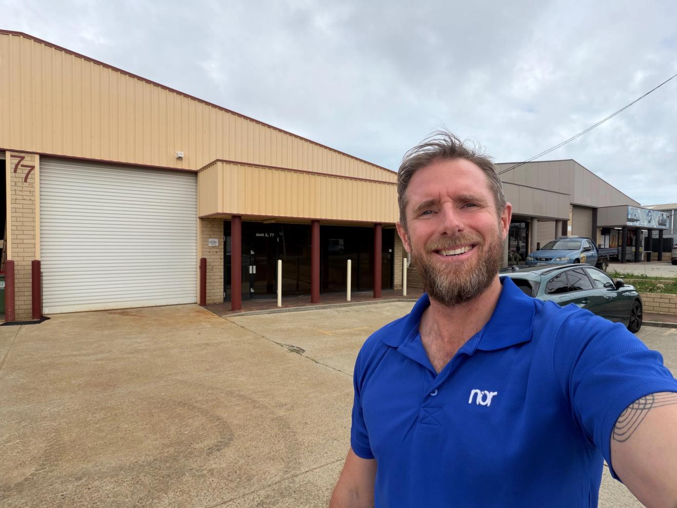 A smiling man in a blue polo shirt with a "nor" logo stands outside a beige commercial building with a large roller door and the number 77.