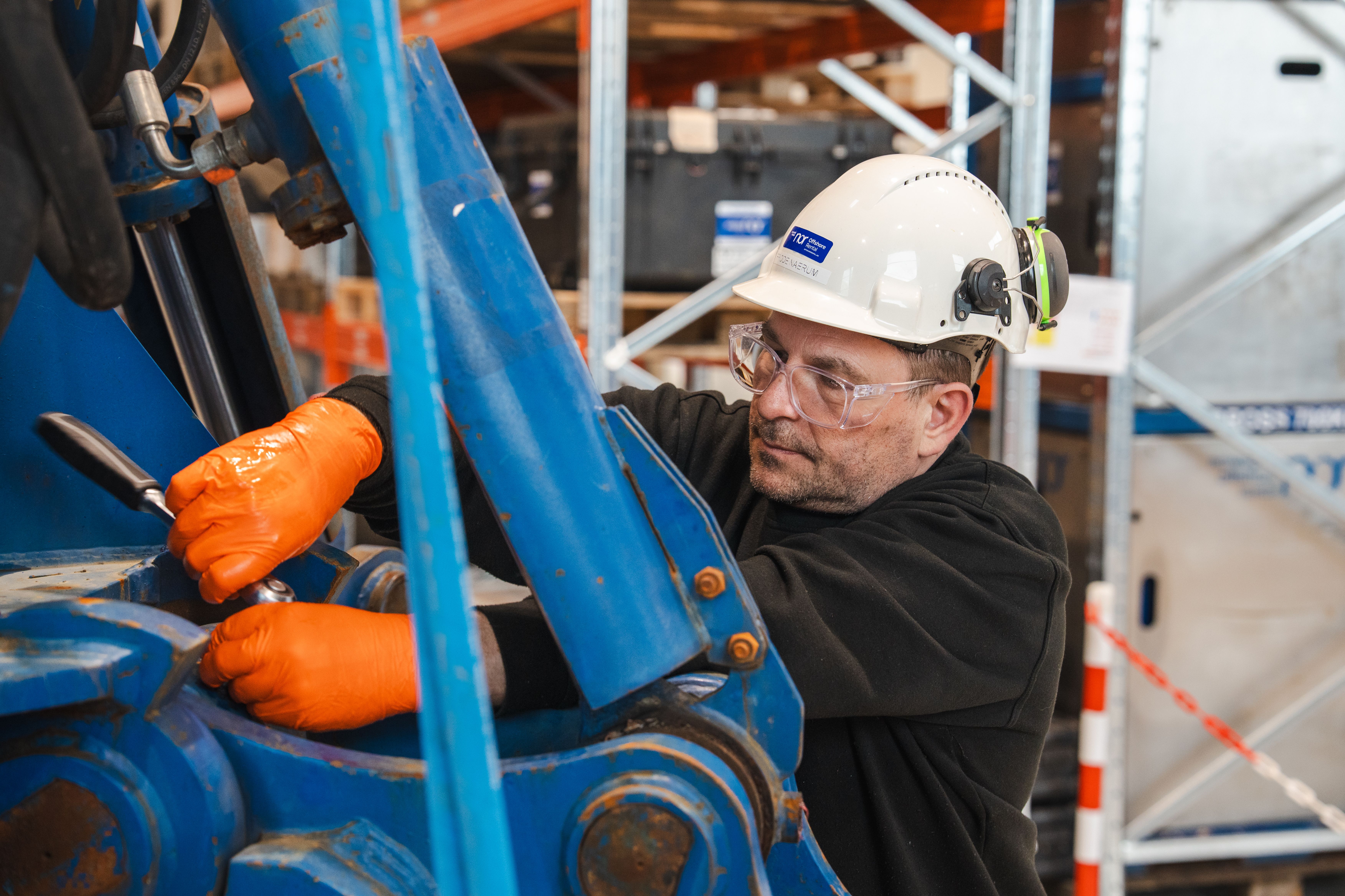 A man in a hard hat, safety glasses, and orange gloves works on large blue machinery.