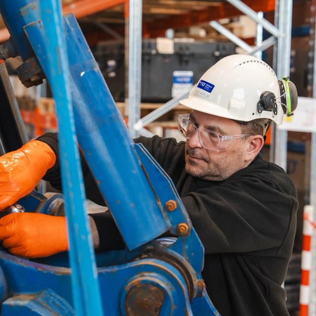 A man in a hard hat and safety glasses, wearing orange gloves, works on blue industrial machinery.