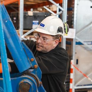 A man in a hard hat, safety glasses, and orange gloves works on blue machinery with a tool.