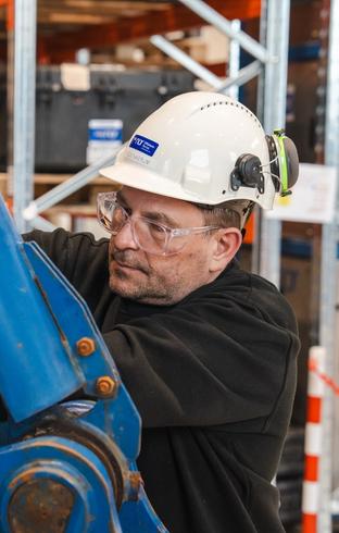 A man in a hard hat, safety glasses, and orange gloves works on large blue machinery.