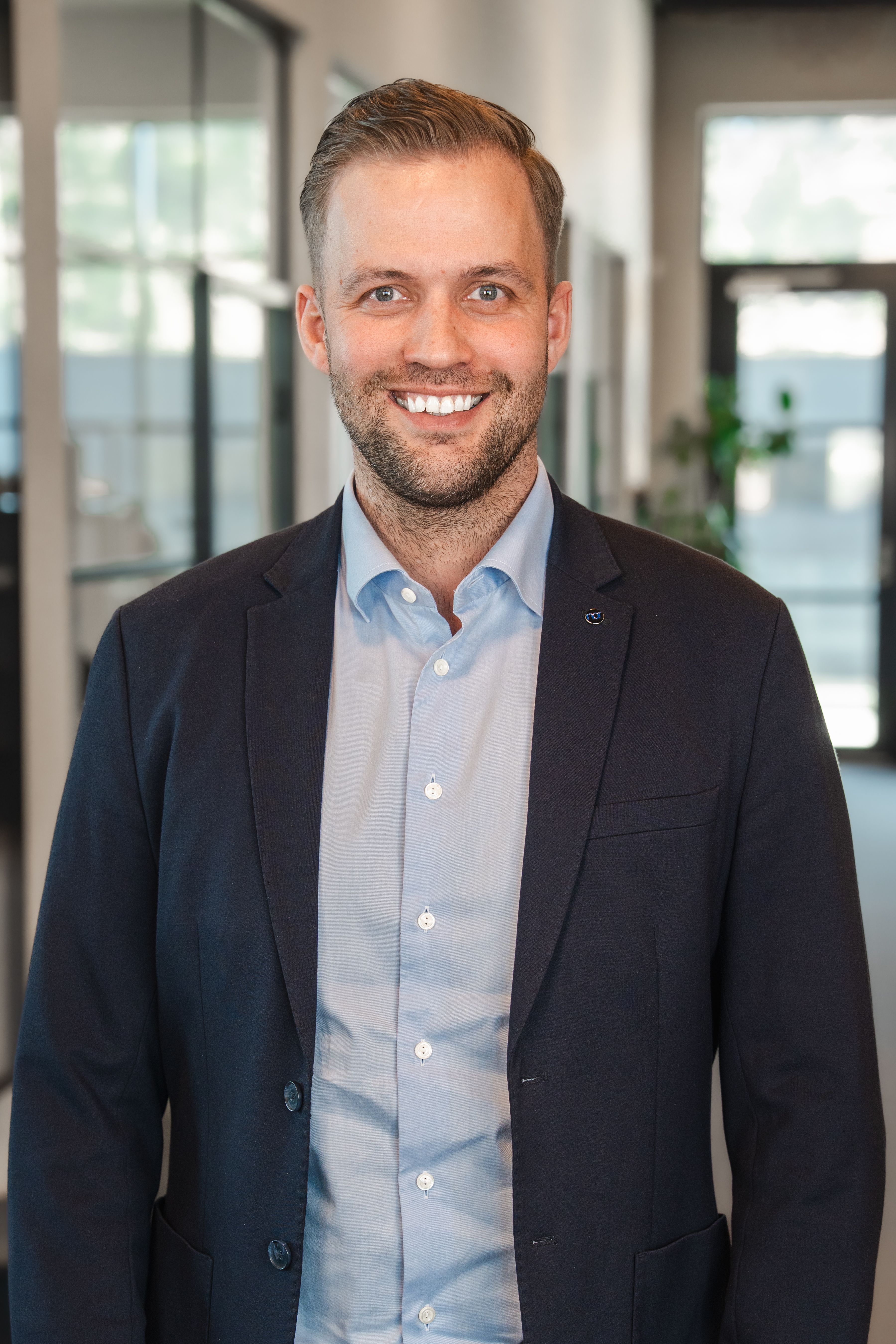 A smiling man with short hair and a beard, wearing a blue shirt and dark blazer, in an office.