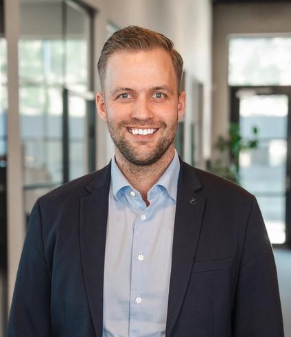 A smiling man with short hair and a beard, wearing a blue shirt and dark blazer, in an office.
