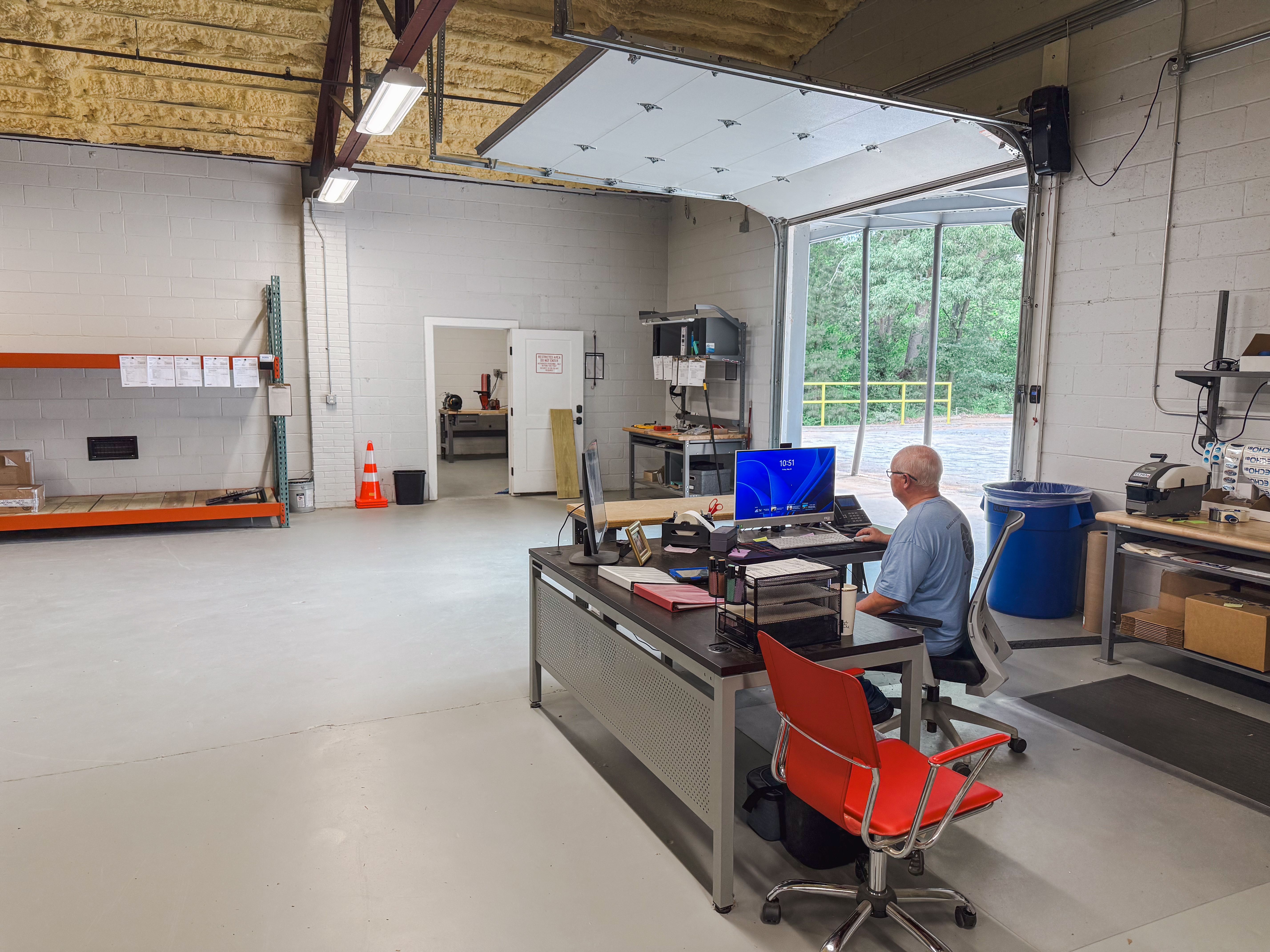 A man works at a desk in a clean workshop with an open garage door leading outside.