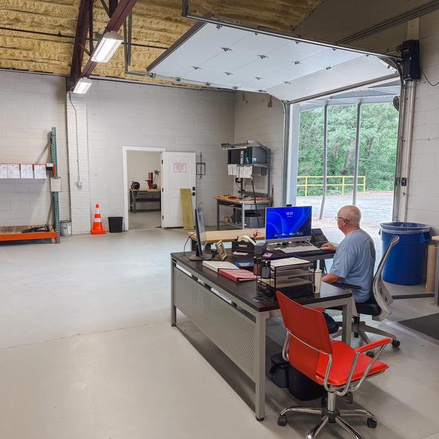 A man works at a desk in a clean workshop with an open garage door leading outside.