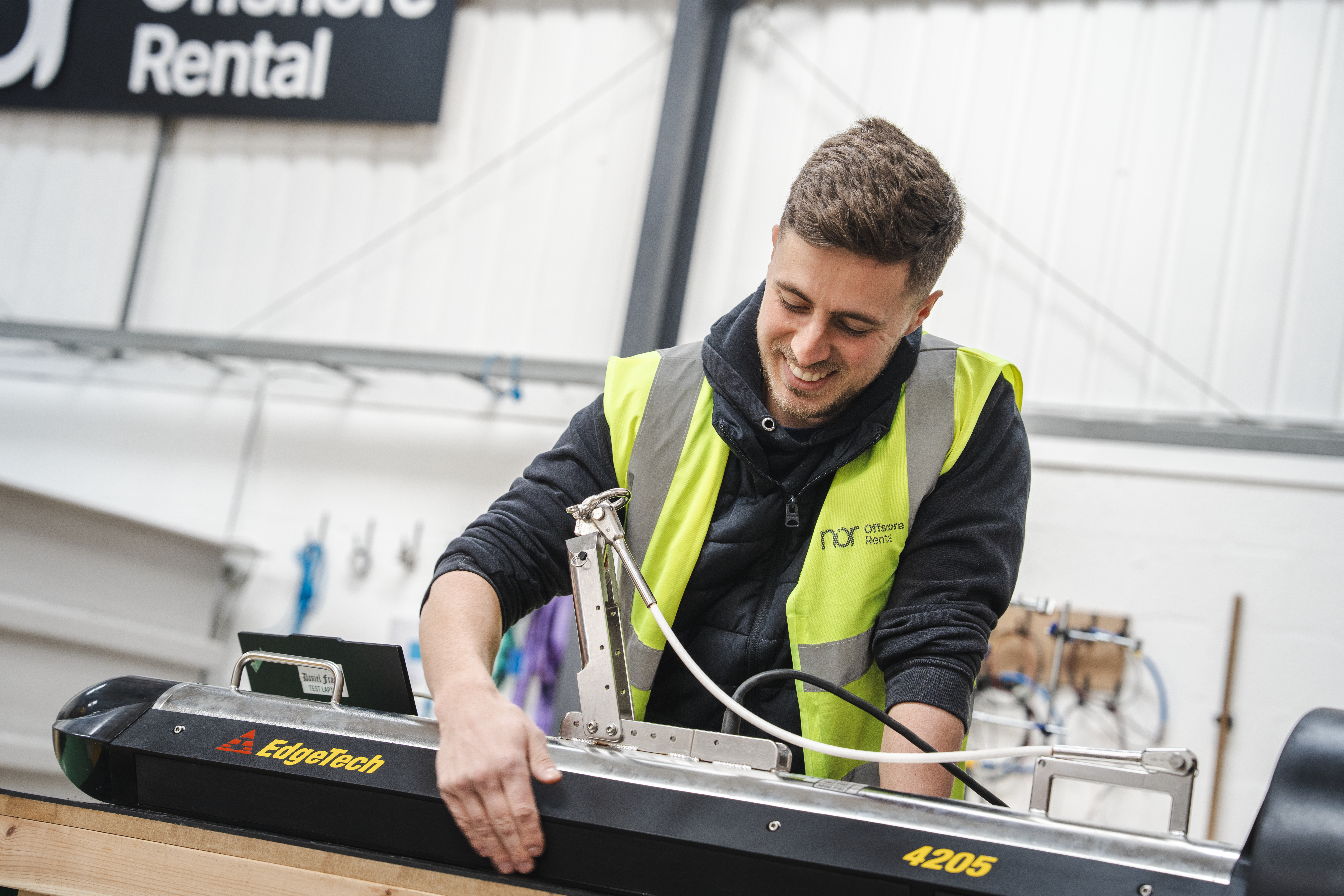 A smiling man in a high-visibility vest works on industrial equipment.