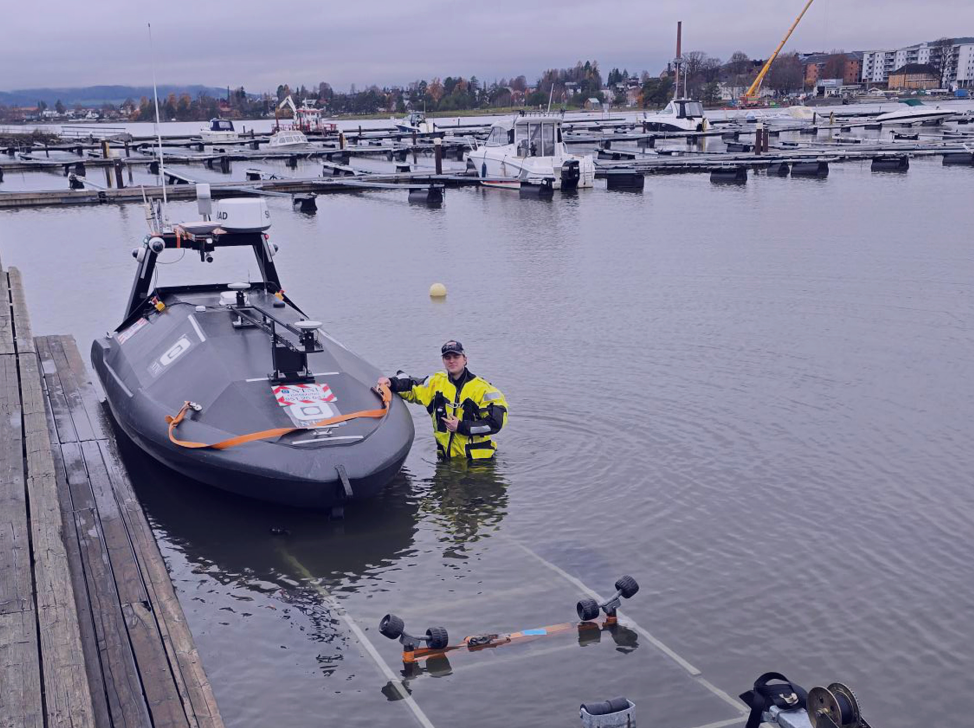 A person in a yellow dry suit stands waist-deep in water next to a dark grey autonomous boat on a boat ramp.