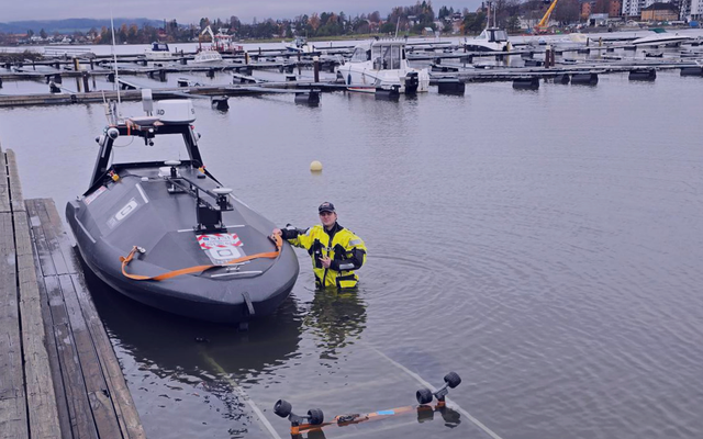 A person in a yellow dry suit stands waist-deep in water next to a dark grey autonomous boat on a boat ramp.