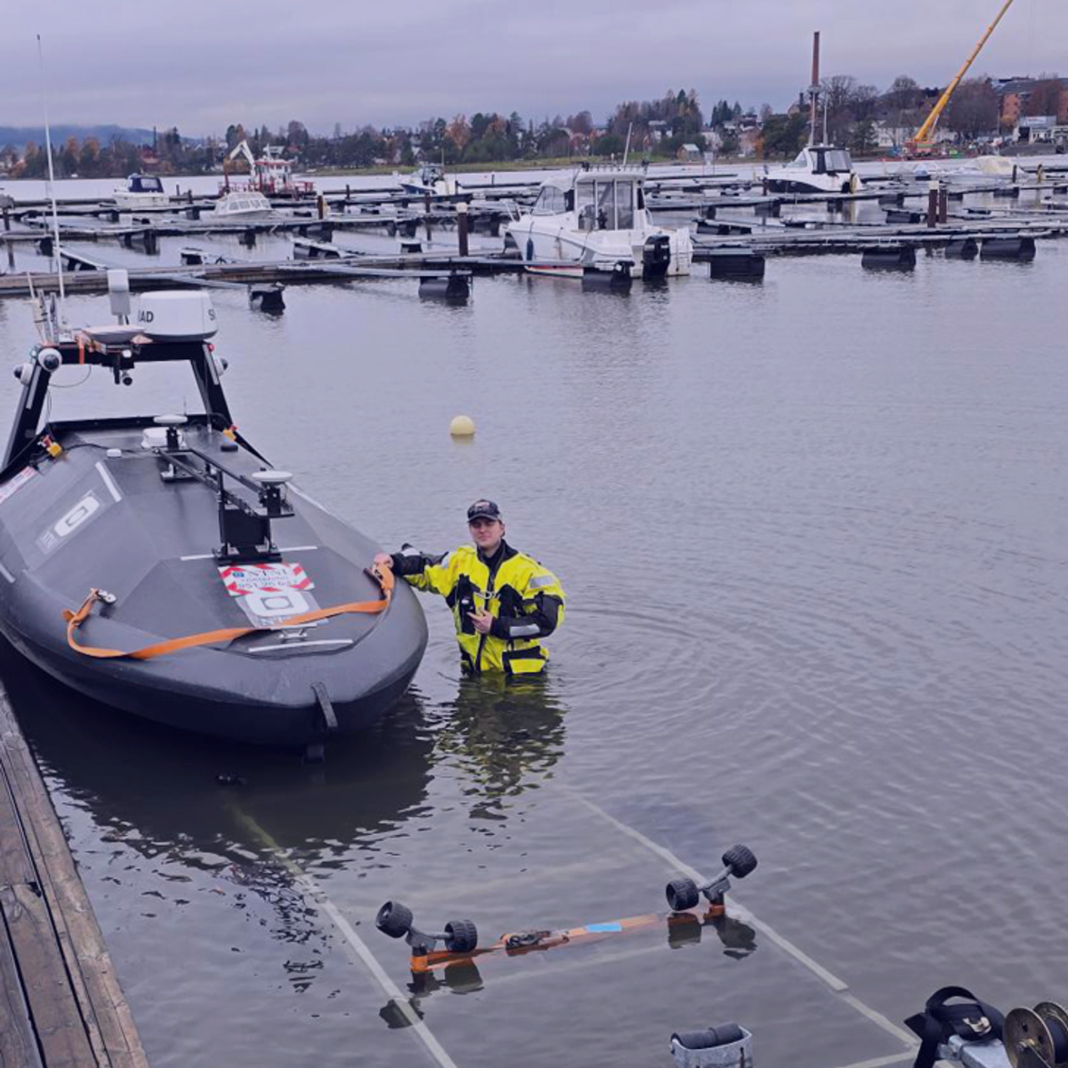 A person in a yellow dry suit stands waist-deep in water next to a dark grey autonomous boat on a boat ramp.