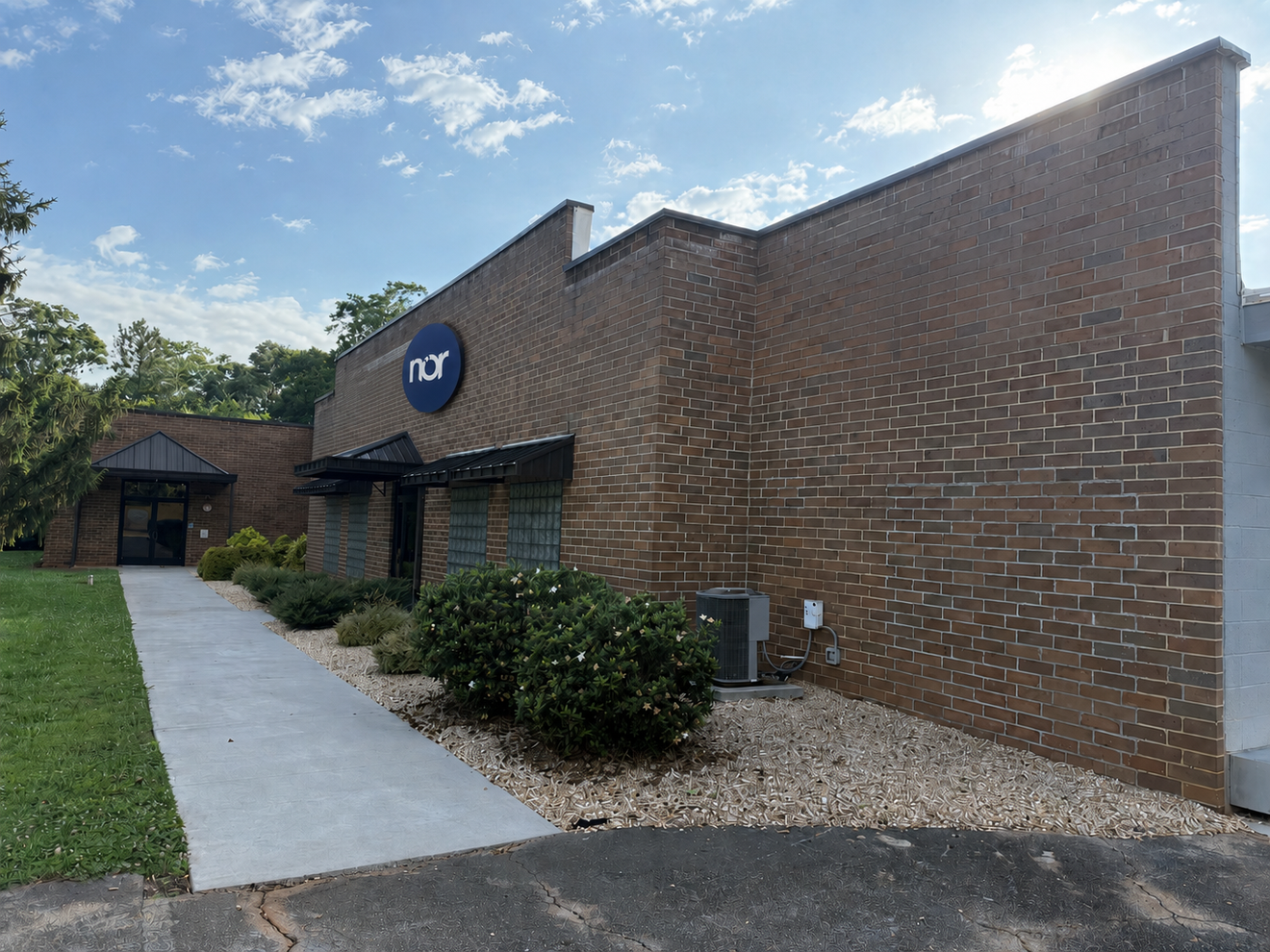 A brick building with a "nor" logo, a walkway, and landscaping under a bright sky.