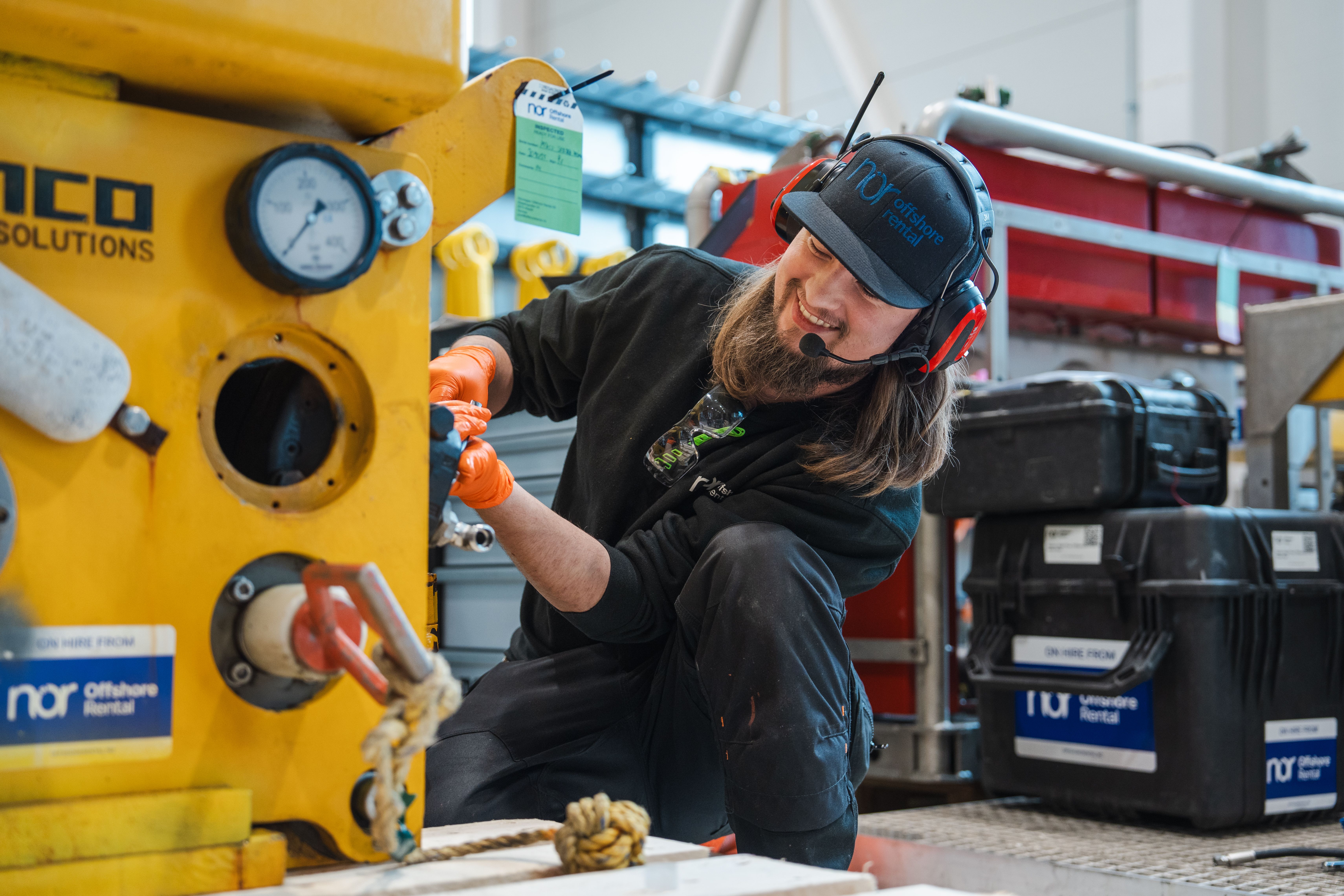 A smiling worker wearing a headset and orange gloves inspects a yellow industrial machine.