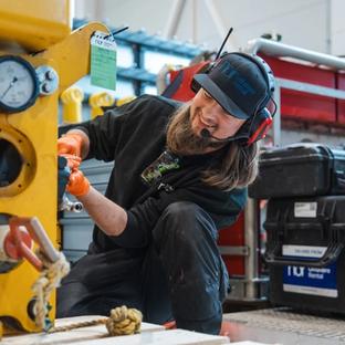 A smiling worker wearing a headset and orange gloves inspects a yellow industrial machine.